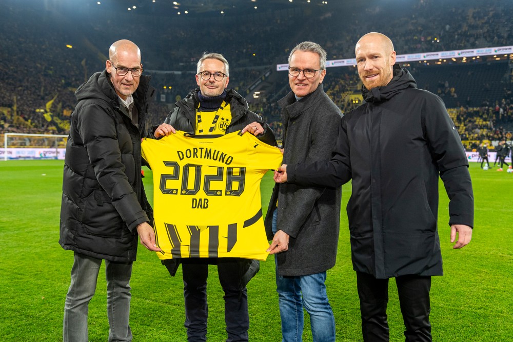 Four men stand on a soccer field, holding a yellow sports jersey with the year 2028 displayed on it.