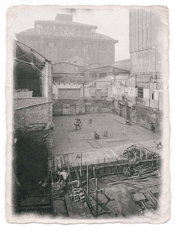 Une photo historique en noir et blanc d'un chantier de construction, avec des ouvriers et des bâtiments en arrière-plan.
