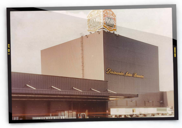 Un grand bâtiment industriel avec un logo en haut et des camions garés près d'un quai de chargement.