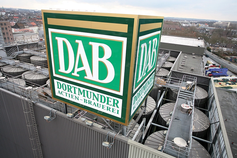Un grand panneau de brasserie vert et blanc est monté sur un toit, surplombant des équipements industriels.