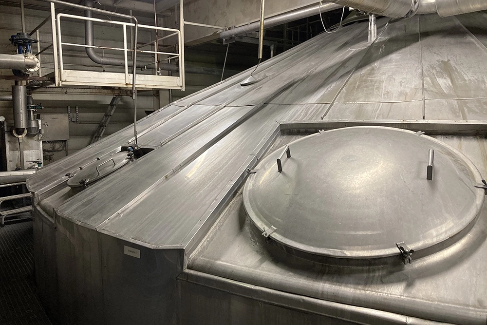 Un grand récipient de brassage en acier inoxydable avec un couvercle en dôme et plusieurs trappes. À l’arrière-plan, des tuyaux et une plateforme en métal.