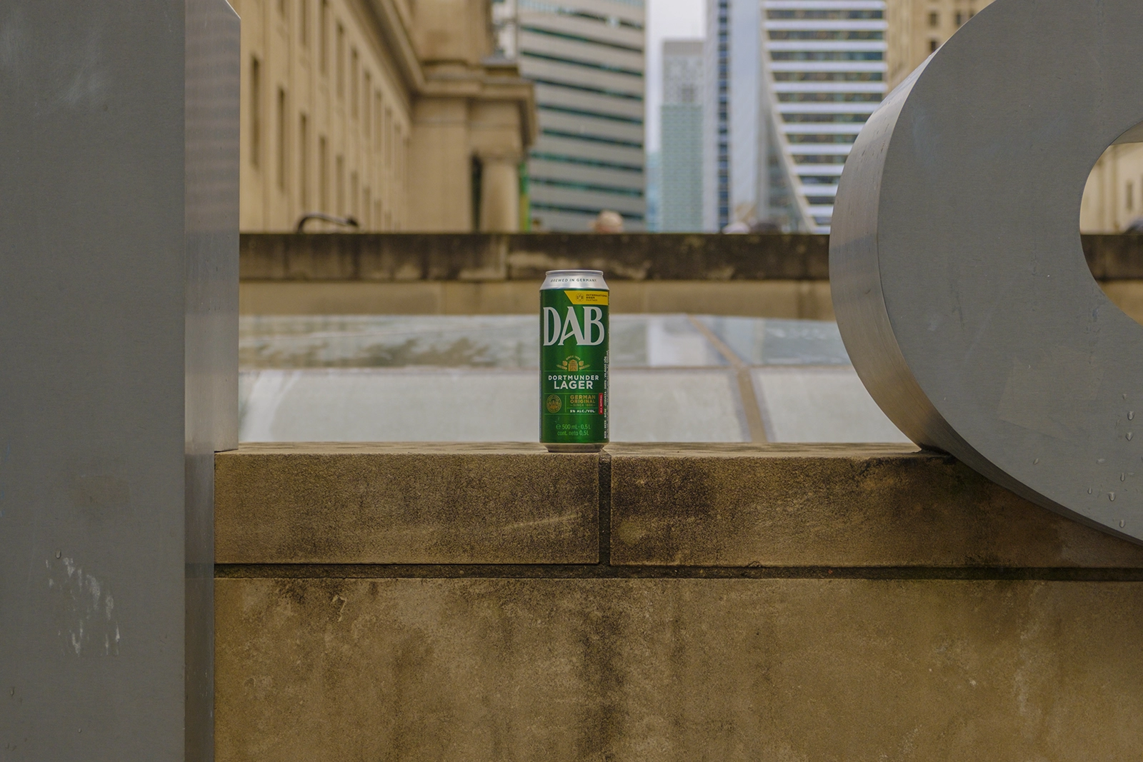 Une canette de bière DAB verte est posée sur une corniche en pierre dans un environnement urbain. De hauts bâtiments modernes s’élèvent en arrière-plan flou.