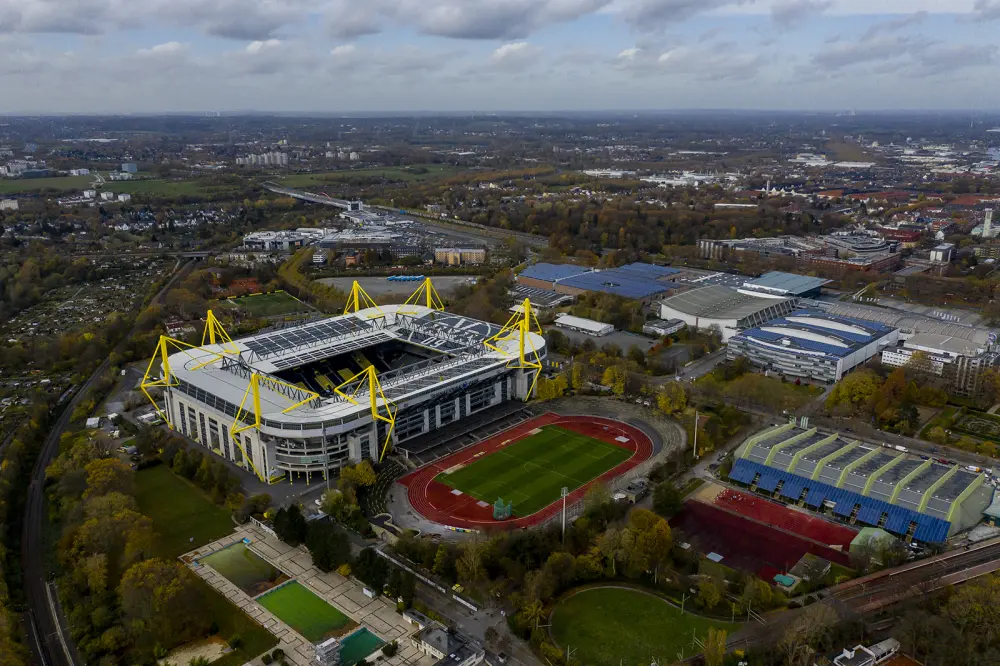 Vue aérienne d’un grand stade avec des supports jaunes distinctifs, entouré d’un complexe sportif et de zones vertes.