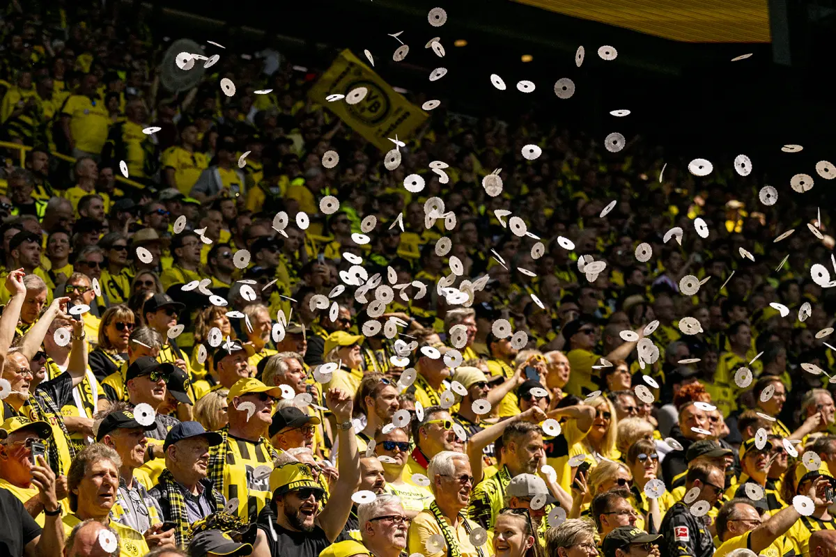 A crowd of excited soccer fans in yellow and black celebrate by tossing paper rolls into the air at a stadium.