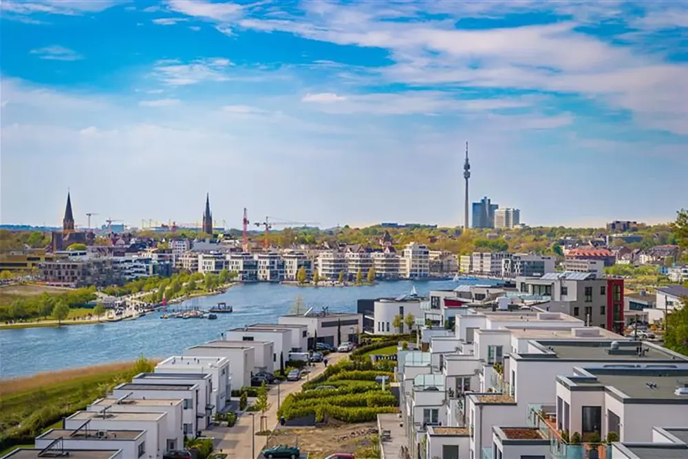 Des immeubles résidentiels modernes bordent un canal avec des chemins et des espaces verts. En arrière-plan se trouvent le centre-ville, des églises et la tour de télévision.