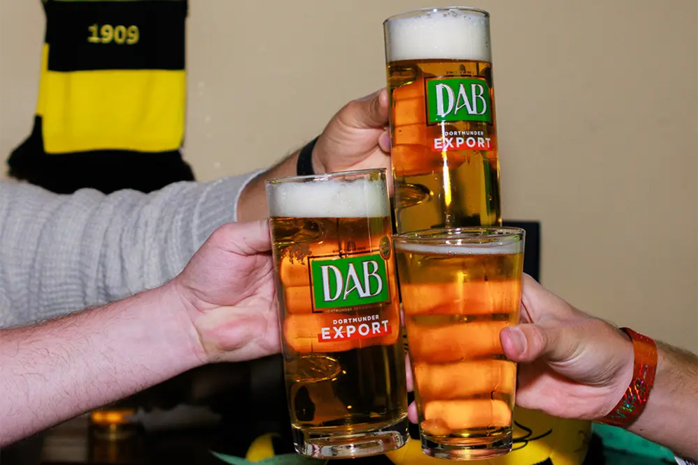Trois personnes trinquent avec des verres remplis de bière ambrée, célébrant ensemble dans une ambiance chaleureuse.