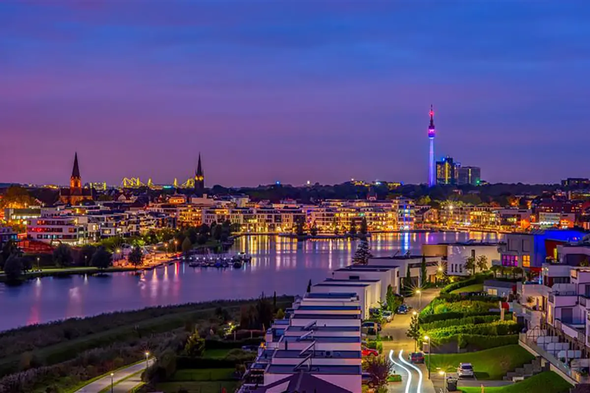 Modern houses line a canal with illuminated streets. In the background, city center, churches and TV tower are lit up.