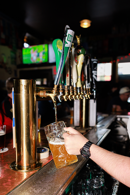 A person is pouring beer from a tap into a clear mug at a bar with dim lighting.
